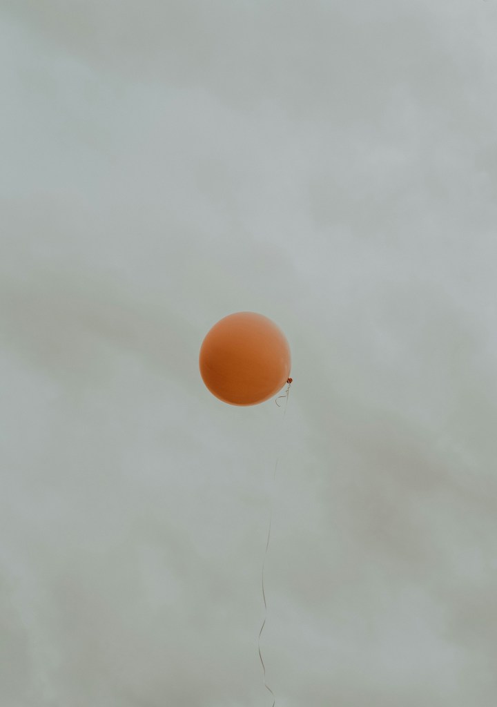 A single orange balloon floating against a cloudy sky.